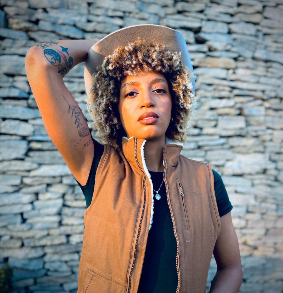 Screenshot 2025-08-06 at 13.48.28 Person with curly hair and tattoos wearing a vest and hat, standing against a stone wall.