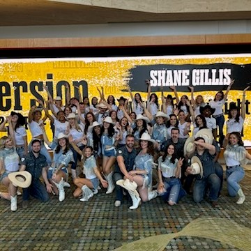 Group of people in cowboy attire posing in front of a 'Shane Gillis' billboard.
