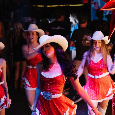 Women in red dresses and cowboy hats dancing in a crowded venue.