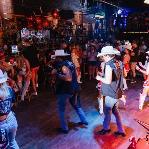 People dancing in a bar wearing cowboy hats and boots, surrounded by a lively crowd.