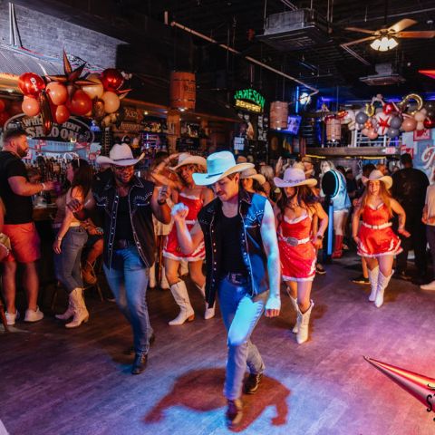 People in cowboy hats line dancing in a bar with balloons and festive decor.