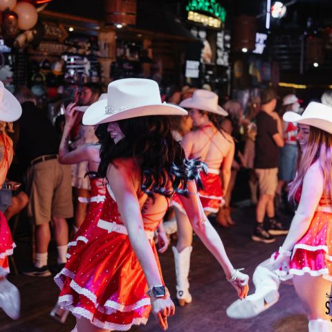 Dancers in red dresses and white hats perform at a lively bar with a crowd watching.
