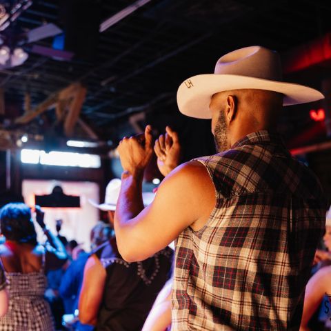 People dancing in a lively bar, wearing cowboy hats and casual clothes, with colorful lighting overhead.