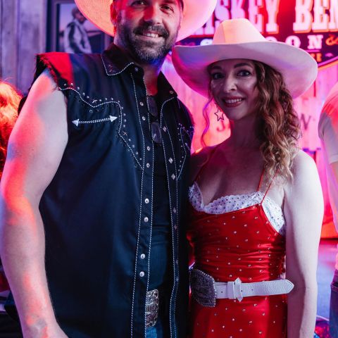 Man and woman in cowboy hats, smiling indoors, with neon sign in background.