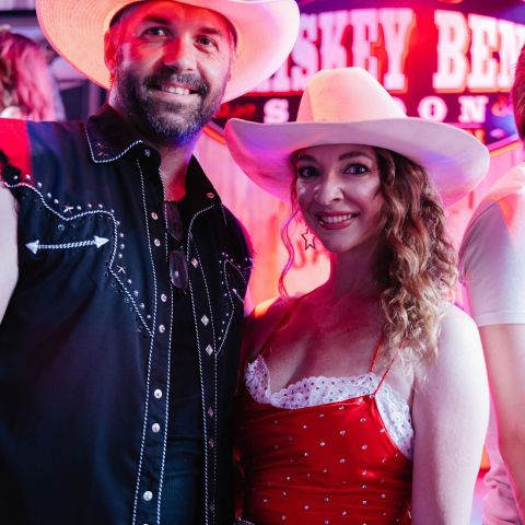Man and woman in cowboy hats and western attire, smiling at a bar with a neon sign.