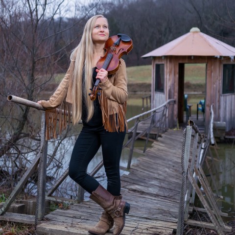 Woman with long hair holding violin on wooden bridge near rustic hut.